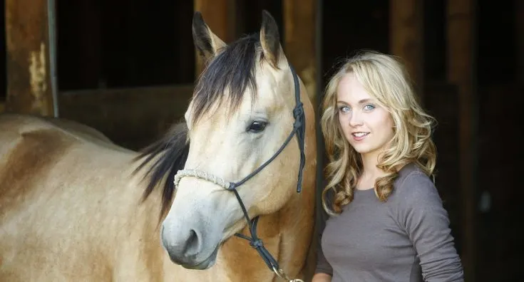 Amber Marshall as Amy Fleming, standing next to a horse in a rustic barn setting